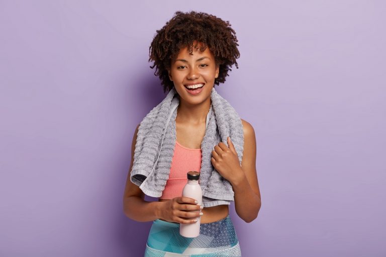 Glad Charming Fitness Woman Drinks Cold Water, Being Thirsty After Running, Has Towel On Neck, Wears Top And Leggings, Stands Against Purple Studio Wall. People, Cardio Training And Healthy Lifestyle