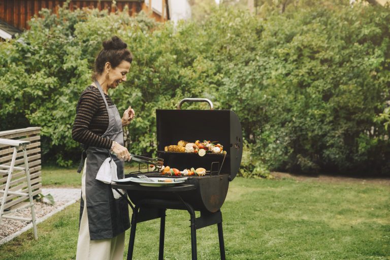 Side View Of Smiling Woman Cooking Dinner On Barbecue Grill At Back Yard During Garden Party